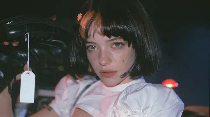 medium close-up of a young Caucasian American woman with pale skin and light freckles, a glossy black blunt jaw-length bob with heavy bangs and one clay-orange streak through the bangs, a small flat clay-orange…