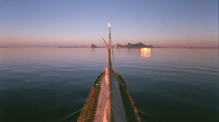 low angle up the mast of a long black ancient Greek galley with a single mast, a square sail and two banks of oars, a small white satellite dish at the top, distant rocks glowing gold on the horizon, dusk, low gold…